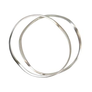May include: Two intertwined silver bangles with a minimalist design. The bracelets are thin, with a slightly irregular shape, and appear to be made of polished metal. The bangles are set against a white background.