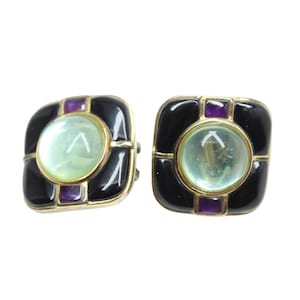 Puede incluir: Un par de pendientes cuadrados de clip. Cada pendiente presenta una piedra central redonda de color verde pálido, rodeada por un marco dorado, esmalte negro y detalles morados. Los pendientes tienen un diseño geométrico vintage.