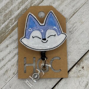 May include: A retractable badge reel featuring a smiling fox face with blue ears and accents, outlined in black. The fox is on a brown tag with the letters "HGC" cut out. The reel has a clear plastic strap.