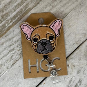 Puede incluir: Una insignia bordada de un bulldog francés marrón y blanco con orejas rosas, unida a un portagafete retráctil de plástico transparente con un clip plateado. El portagafete está unido a una tarjeta marrón con el texto "HGC" impreso en ella.