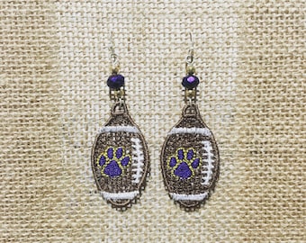 May include: A pair of brown and white football-shaped earrings with a purple paw print design. The earrings have a purple and gold bead at the top.