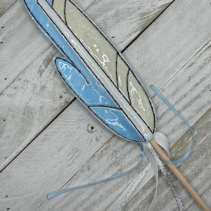 Può includere: Una piuma blu e bianca con un bastone di legno e nastri blu e argentati attaccati.