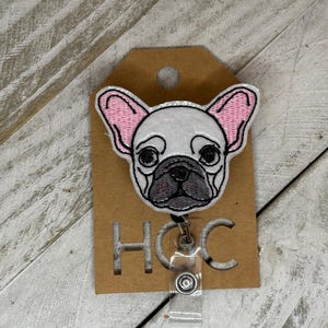 Puede incluir: Un portatarjetas con un diseño bordado de bulldog francés. El perro tiene la cara blanca, las orejas rosas y la nariz negra. El portatarjetas está sujeto a una etiqueta marrón con las letras "HGC" recortadas.