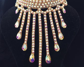 May include: A dazzling choker necklace with a tan leather base, adorned with numerous iridescent rhinestones. The design features a wide collar and cascading strands of rhinestones, culminating in large teardrop-shaped gems. The necklace is displayed on a black mannequin.