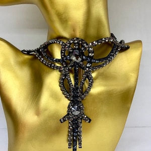Op de afbeelding: Een zwarte choker met strass en een hangende hanger. De ketting is gemaakt van kleine, gefacetteerde strasssteentjes die in een metalen frame zijn gezet. De hanger is druppelvormig en is ook gemaakt van strass.