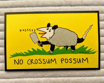 May include: A yellow sticker with a cartoon possum holding a knife and saying "HISSsssssss". The possum is standing on green grass and the sticker says "NO CROSSUM POSSUM".