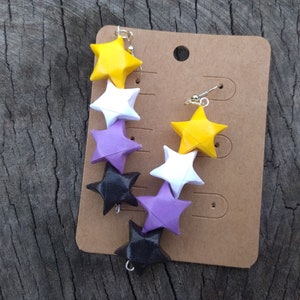 Peut inclure: Une paire de boucles d'oreilles avec une chaîne et six étoiles en origami. Les étoiles sont jaunes, blanches, violettes et noires. Les boucles d'oreilles sont présentées sur une carte marron.