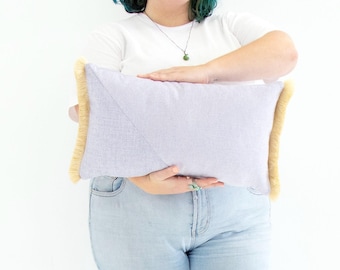 Funda de cojín rectangular texturizada en tonos lila, morado y amarillo con flecos de algodón