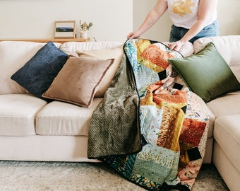 May include: A white couch with a patchwork quilt draped over it. The quilt has a variety of colors and patterns, including red, orange, yellow, green, blue, and brown. There are also three throw pillows on the couch, one navy blue, one brown, and one green.