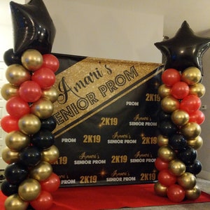 K&ouml;nnte beinhalten: Ein schwarzes und goldenes Banner mit dem Text "Amari's Senior Prom 2K19" und einem roten Teppich davor. Zwei Ballons&auml;ulen, eine auf jeder Seite des Banners, bestehen aus schwarzen, goldenen und roten Ballons. Jede S&auml;ule hat einen gro&szlig;en schwarzen sternf&ouml;rmigen Ballon oben.