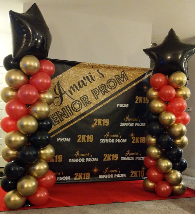 K&ouml;nnte beinhalten: Ein schwarzes und goldenes Banner mit dem Text "Amari's Senior Prom 2K19" und einem roten Teppich davor. Zwei Ballons&auml;ulen, eine auf jeder Seite des Banners, bestehen aus schwarzen, goldenen und roten Ballons. Jede S&auml;ule hat einen gro&szlig;en schwarzen sternf&ouml;rmigen Ballon oben.