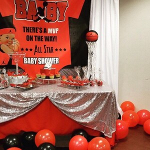 May include: A red and black baby shower banner with the text "BA-BY", "THERE'S A MVP ON THE WAY!" and "*** ALL STAR *** BABY SHOWER". A basketball is in a hoop on a silver stand. A table is covered with a silver sequin tablecloth and red tablecloth. There are cupcakes and other desserts on the table. There are red and black balloons on the floor.