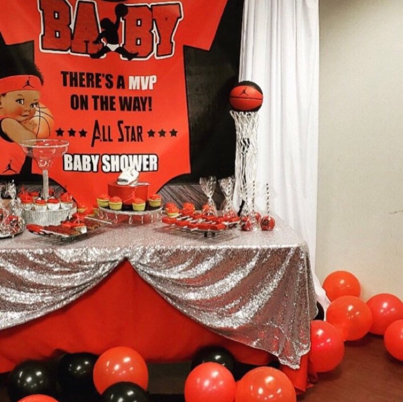 May include: A red and black baby shower banner with the text "BA-BY", "THERE'S A MVP ON THE WAY!" and "*** ALL STAR *** BABY SHOWER". A basketball is in a hoop on a silver stand. A table is covered with a silver sequin tablecloth and red tablecloth. There are cupcakes and other desserts on the table. There are red and black balloons on the floor.