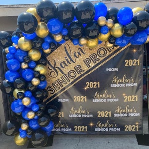 K&ouml;nnte beinhalten: Ein schwarzes Banner mit goldenen Glitzerbuchstaben, auf dem "Kailen Senior Prom 2021" steht. Das Banner ist mit blauen, schwarzen und goldenen Luftballons geschm&uuml;ckt.