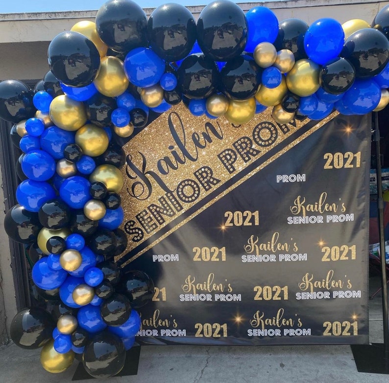 K&ouml;nnte beinhalten: Ein schwarzes Banner mit goldenen Glitzerbuchstaben, auf dem "Kailen Senior Prom 2021" steht. Das Banner ist mit blauen, schwarzen und goldenen Luftballons geschm&uuml;ckt.