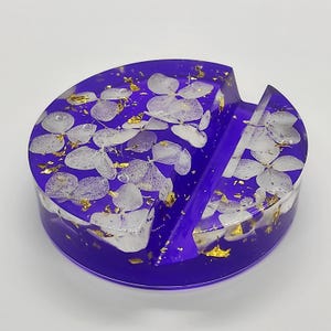 May include: A vibrant purple resin phone stand with a circular design. The stand is embedded with white floral elements and gold leaf flakes. It features a slot to hold a phone upright, offering a decorative and functional accessory.