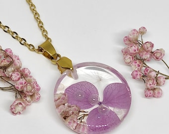 May include: A round resin pendant necklace featuring embedded purple hydrangea petals and small pink flowers. The pendant is attached to a gold-toned metal chain. The design includes clear resin with visible botanical elements and a metallic bail.