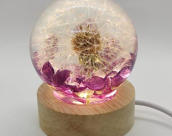 May include: A decorative lamp with a clear resin sphere containing a dandelion and purple petals. The sphere rests on a round, light-coloured wooden base. The lamp emits a soft, warm glow and has a white power cord.