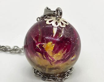May include: A spherical pendant necklace featuring a clear resin orb containing a preserved magenta and yellow flower. The orb is topped with a silver-toned metal cap and a small loop, attached to a silver-toned metal chain that rests at the base.