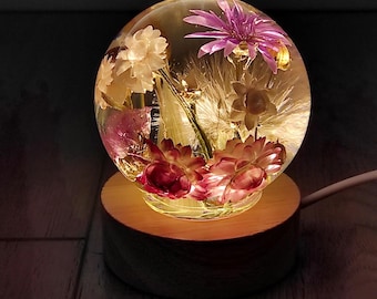 May include: A clear glass sphere with dried flowers encased in resin, illuminated from below by a warm light. The sphere sits on a wooden base.