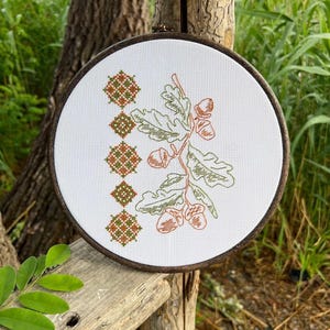 May include: A cross-stitch embroidery hoop featuring an oak branch with acorns in shades of brown and green. The design is set against a white fabric, with a series of diamond-shaped patterns on the left side. The wooden hoop is visible.