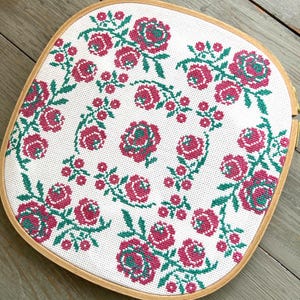May include: A square wooden embroidery hoop containing white Aida fabric with a cross-stitch pattern of repeating pink and green rose motifs. The floral design features clusters of roses and small buds arranged in a symmetrical grid across the fabric.
