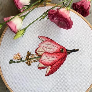Peut inclure: Un gros plan d'une broderie au point de croix d'une fleur rose avec une longue tige fine et des feuilles. La broderie est encadrée dans un cercle en bois.