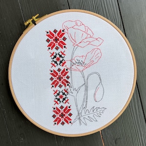 Może przedstawiać: Biały tamborek do haftu z haftem krzyżykowym przedstawiającym czerwone i czarne maki oraz czerwony i czarny wzór geometryczny.