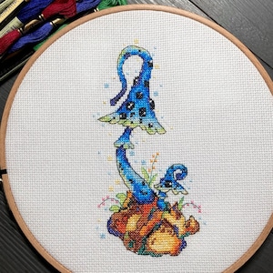 May include: Cross-stitch embroidery of a blue and green mushroom with black spots, surrounded by yellow and orange flowers on a white background.
