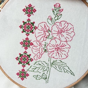 May include: A close-up of a hand-stitched embroidery hoop featuring a floral design. The design includes pink flowers with green leaves and stems. The flowers are outlined in red thread and the leaves are outlined in green thread. The background is a white fabric.