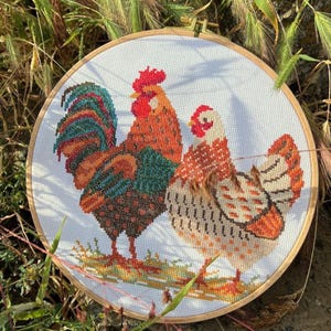 May include: A circular wooden embroidery hoop containing a cross-stitch pattern of a rooster and a hen. The rooster features vibrant red, orange, blue, and dark brown plumage. The hen is depicted in lighter shades of orange, white, and brown.