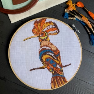 May include: A colourful cross-stitch embroidery of a hoopoe bird with a long beak and a distinctive crest of feathers. The bird is stitched in shades of orange, yellow, blue, and brown on a white canvas.