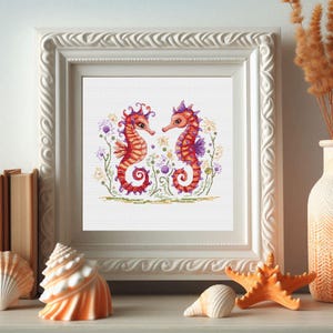 Puede incluir: Una obra de arte en punto de cruz enmarcada con dos caballitos de mar naranjas y rojos enfrentados, rodeados de flores moradas y amarillas. La obra está en un marco blanco adornado, expuesta en una estantería con conchas y una estrella de mar.