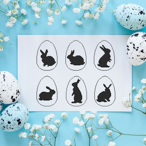 May include: A white sheet of paper with six black bunny silhouettes inside egg shapes. The paper is surrounded by white baby's breath flowers on a light blue background.