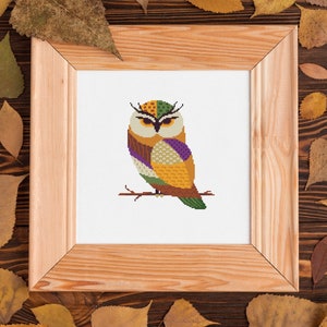 Puede incluir: Un bordado de punto de cruz enmarcado de un búho colorido posado en una rama. El búho presenta patrones geométricos en tonos amarillos, verdes, morados y marrones. El marco de madera clara está rodeado de hojas de otoño.