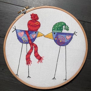 Op de afbeelding: Een borduurring met kruissteek met twee grillige vogels. De vogels hebben blauwe lichamen, gele snavels en lange, dunne zwarte poten. Eén vogel draagt een rode sjaal en hoed, de andere een groene hoed. De vogels kussen elkaar.