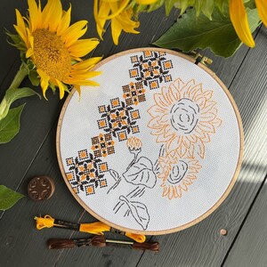 May include: A white embroidery hoop with a cross-stitch design featuring black and orange sunflowers and geometric patterns. The hoop is on a dark gray surface.