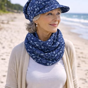 Pode incluir: Um chapéu azul marinho com aba e um cachecol infinito a condizer, ambos adornados com um padrão floral branco. O chapéu e o cachecol são usados com uma camisa branca e um cardigan bege. A cena passa-se numa praia.