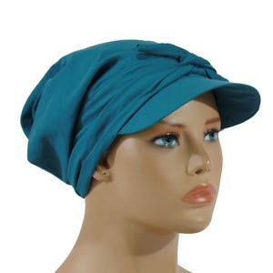 Pode incluir: Chapéu azul-petróleo com viseira. O chapéu tem um design estilo turbante com um detalhe de nó na parte superior. O tecido parece macio e drapeja ao redor da cabeça. A viseira tem uma forma curva.