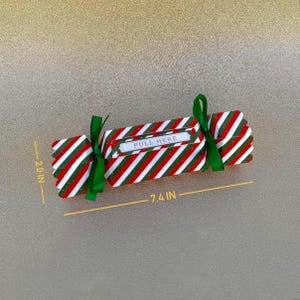 May include: A candy-shaped paper box with red, white, and green diagonal stripes, tied with green ribbons at both ends. The center features a white label with the text PULL HERE. The box measures 7.4 inches in length and 2.0 inches in height.