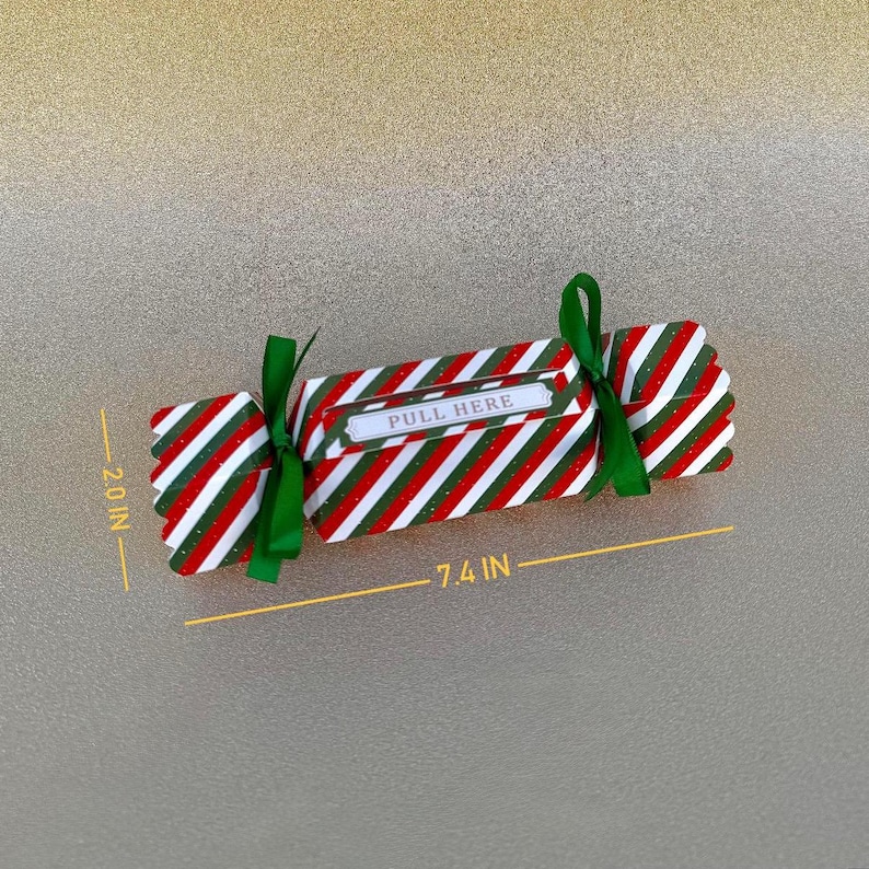 May include: A candy-shaped paper box with red, white, and green diagonal stripes, tied with green ribbons at both ends. The center features a white label with the text PULL HERE. The box measures 7.4 inches in length and 2.0 inches in height.