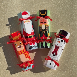 May include: A collection of Christmas crackers with festive designs. The crackers feature illustrations of Santa Claus, a nutcracker, a reindeer, and a snowman, each tied with a red ribbon. The crackers are on a gold glitter surface.