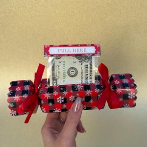 May include: A rectangular gift box featuring a red and black buffalo plaid pattern with white snowflake accents. The box has red ribbon ties on both ends and a top tab labeled PULL HERE, revealing a one-dollar bill inside a clear plastic sleeve.