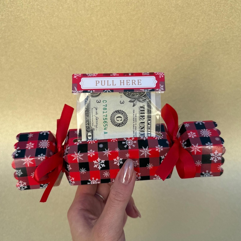 May include: A rectangular gift box featuring a red and black buffalo plaid pattern with white snowflake accents. The box has red ribbon ties on both ends and a top tab labeled PULL HERE, revealing a one-dollar bill inside a clear plastic sleeve.