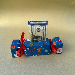 May include: A blue Christmas cracker with red ribbon ties, decorated with festive icons like a snowman, Santa, and gingerbread men. A dollar bill is visible inside, with a "PULL HERE" label.