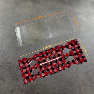 May include: A clear rectangular plastic sleeve measuring 7.75 inches by 3.1 inches and a red and black buffalo plaid cardstock cutout with white snowflake patterns, measuring 8.75 inches by 3.5 inches, featuring a central horizontal slit.