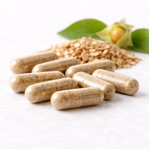 May include: Close-up of several brown capsules, likely supplements, arranged in the foreground. Behind them, a pile of small, tan-colored seeds or grains, with a green leaf and a small, yellow-orange fruit in the background. The capsules are the primary focus.