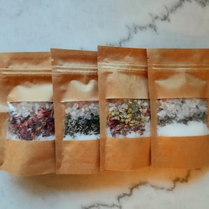 Puede incluir: Cuatro bolsas de papel marrón llenas de sales de baño y flores secas. Las bolsas tienen una ventana transparente que muestra el contenido, incluyendo pétalos de rosa rosas, hierbas verdes y cristales de sal blancos.