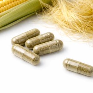 Könnte beinhalten: Nahaufnahme von mehreren grünen Kapseln, wahrscheinlich Nahrungsergänzungsmitteln, auf einer weißen Oberfläche. Im Hintergrund ein frischer Maiskolben mit grünen Blättern und Maisbart. Die Kapseln sind mit einem hellgrünen Pulver gefüllt.