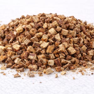 May include: A pile of chopped, dried, and roughly cut pieces of brown and tan herbs. The pieces have a rough, textured appearance and vary in size. The herbs are on a white surface. The image is well-lit, highlighting the natural colors and textures.